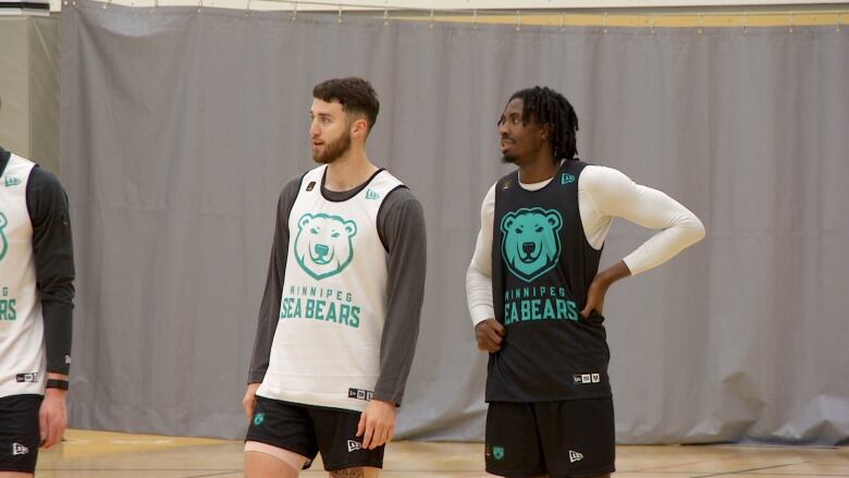 A white man with a white basketball jersey with green lettering stands next to a black man with a black jersey with green lettering. 