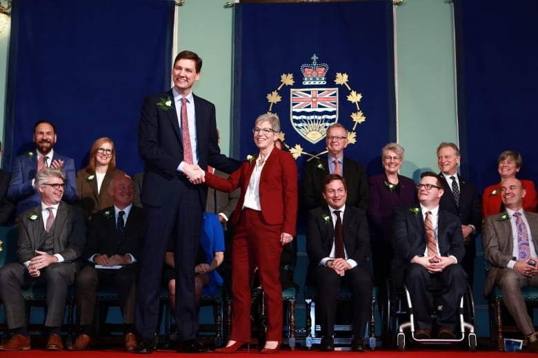 A tall white man shakes the hand of a smaller white woman while other people flank them at an inauguration ceremony.