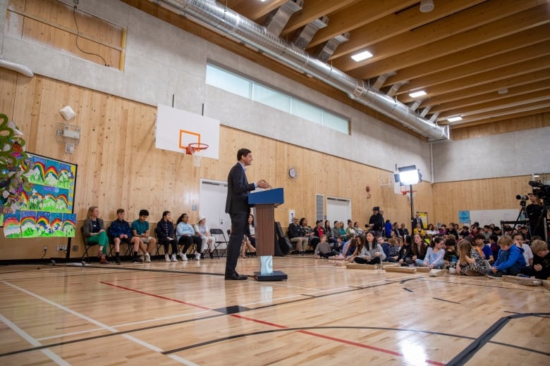 A white man delivers a press conference in a school gymnasium, surrounded by students.