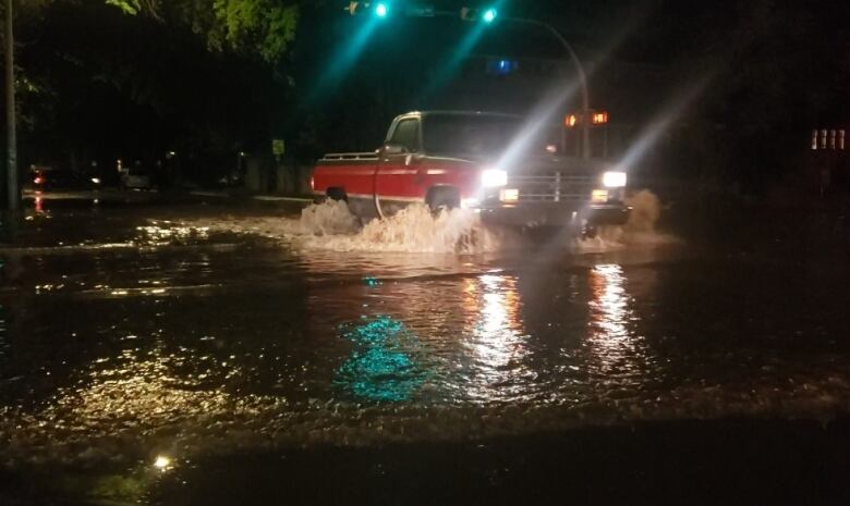 A grey-and-red truck is driving through a large pool of water at an intersection, at nighttime.