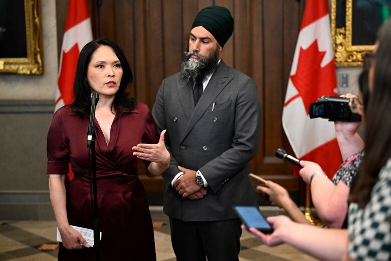 A woman with dark hair and dark red shirt answers questions from reporters. A man in a gray suit looks on.