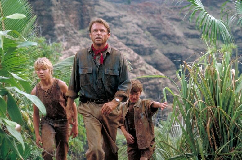 Still frame from the original Jurassic Park movie. Joseph Mazzello, Sam Neill, and Ariana Richards wander through the grounds of Jurassic Park, dirtied and wearing safari clothes.