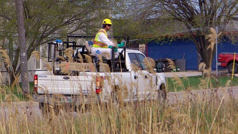 A man wearing protective gear is sitting in the back of a pickup truck, holding a spraying wand which is spraying something into a grassy area.