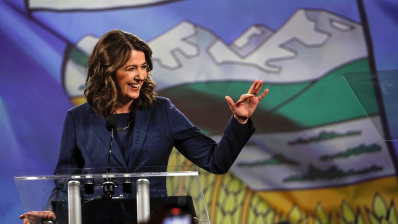 Newly elected premier Danielle Smith waves to supporters from a podium with the Alberta flag flying in the background.