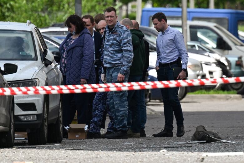 Several people, including one in military fatigues, are shown standing on a road behind police tape. There appears to be debris on the street.