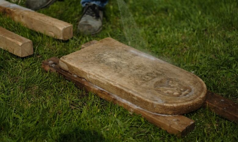 A gravestone sits flat on the grass, supported by wooden planks. 