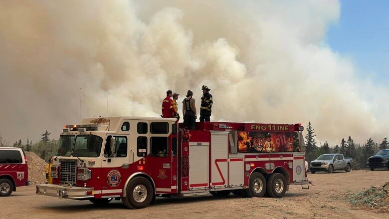 Four people in firefighting gear stand on top of a fire engine with a huge plume of smoke in the background.