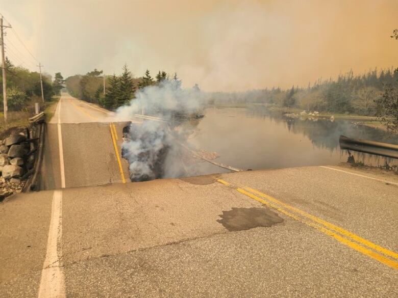 A road is broken and collapsed next to a body of water, with smoke rising from underneath.