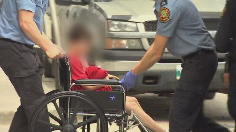 A child is seen on a wheelchair being moved around by emergency responders. The child's face is blurred out.