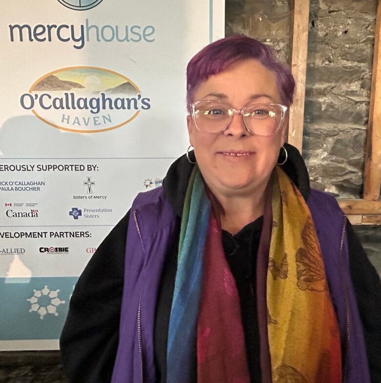 A woman with short purple hair and a multi-coloured scarf stand in front of a white sign and a stone wall. 