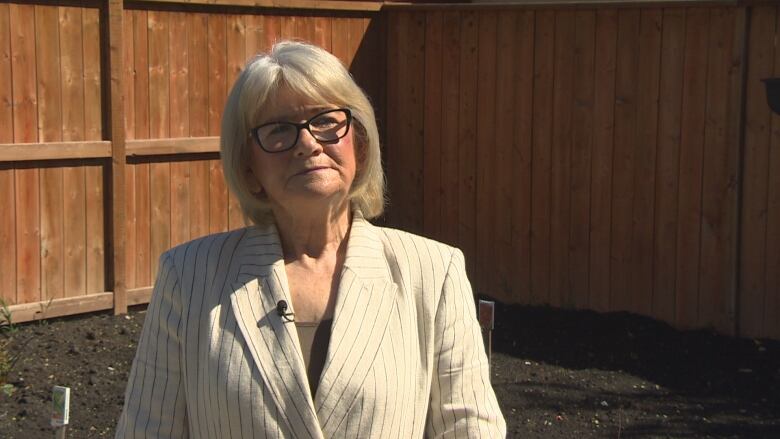 A woman with blonde hair, wearing a cream coloured striped blazer and black glasses. 