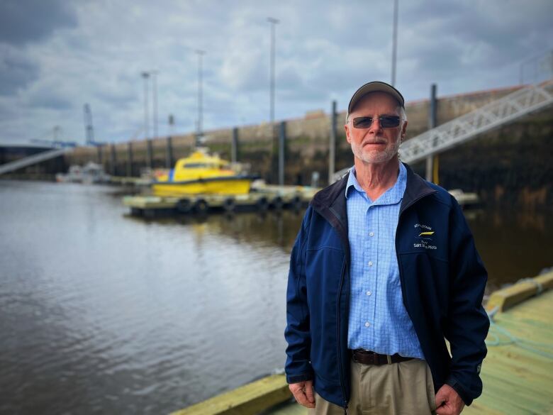 A grey-bearded former marine pilor stands at the pilot dock by the yellow pilot boats in the Saint John harbour wearing dark glasses and a baseball hat. 