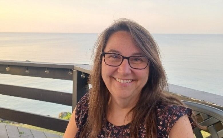 A person smiles at the camera. They are sitting on a deck or patio with a body of water behind them