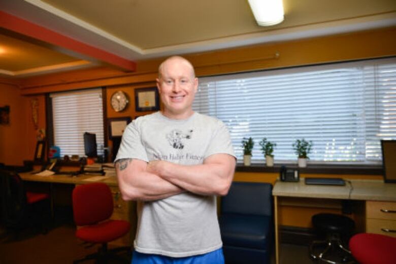 A bald man in a grey T-shirt and blue athletic shorts crosses his arms and smiles for a photo.