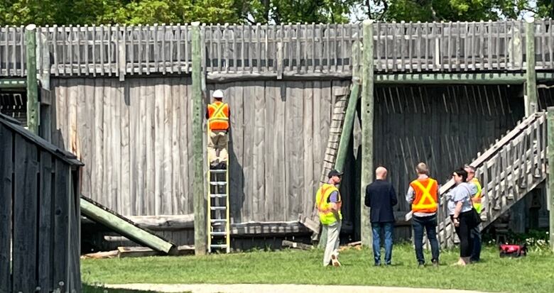 Crew workers are seen standing near a wooden structure. One is leaning against it on a ladder.