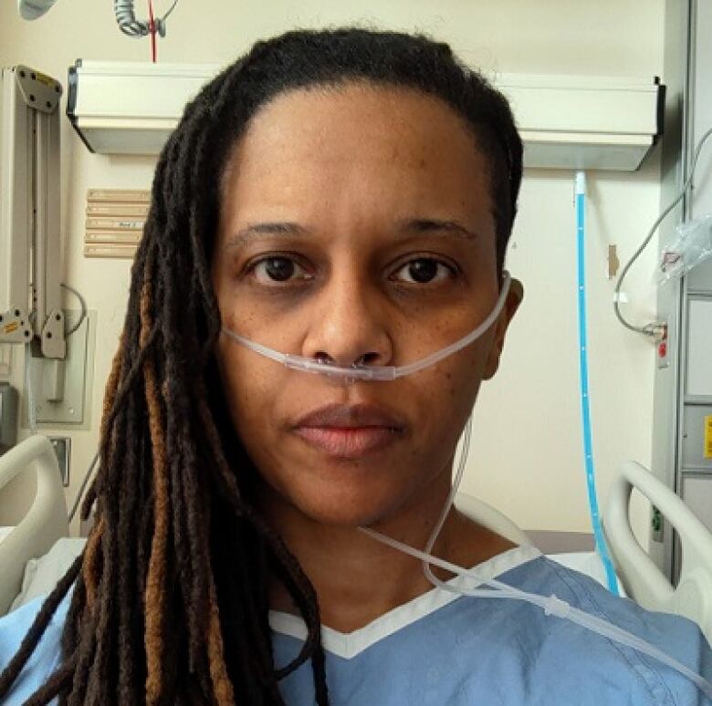 A woman in a blue hospital gown, with an oxygen tube in her nose, is shown in a selfie taken from a hospital bed.