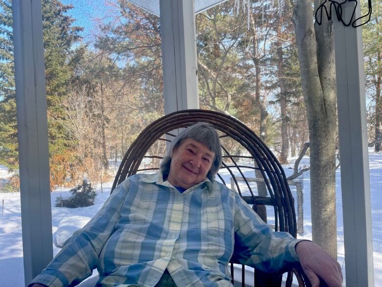 A woman in a plaid shirt sits in a chair in front of a window, with snow and trees in the background. 