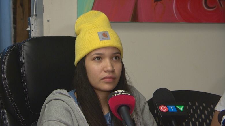 A young woman with long black hair is wearing a yellow Carhartt-brand toque, a grey zip-up hoodie and a blue shirt. She is sitting in a black desk chair, in front of two microphones.