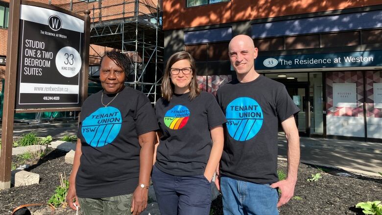 Three people stand wearing shirts that say Tenant Union, they are in front of a sign advertising their building.