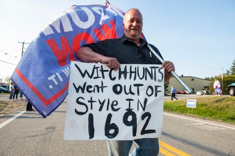 A man holds a flag that says 'Witch hunts went out of style in 1692' while standing in the middle of a road.