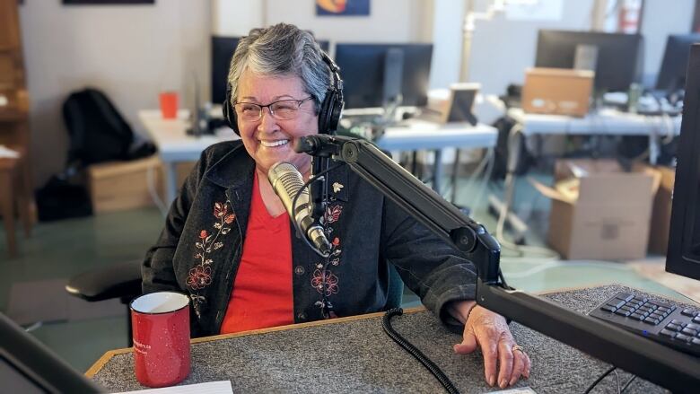 A woman with short grey hair and glasses smiles while being interviewed in a radio studio.