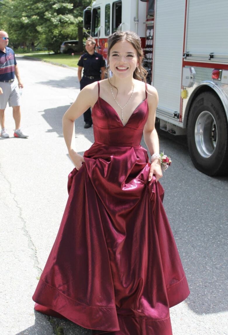 A girl with brown hair smiles in her prom dress. It is a long, burgundy, sleeveless ballgown. 