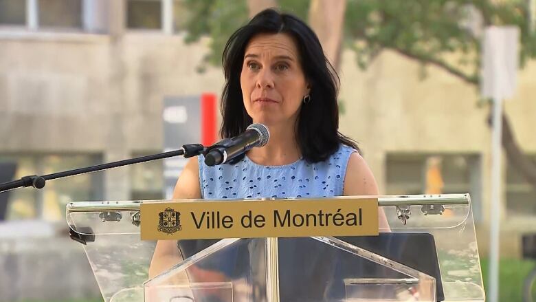 Montreal Mayor Valrie Plante stands at a clear city podium at an outdoor news conference on a hot sunny day, wearing a light blue top.