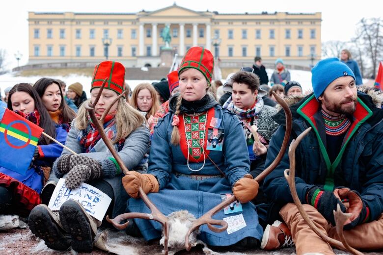 A group of protesters, some wearing red and green hats, sit on the ground outside a legislative building in the winter. A woman at the centre of the frame holds large reindeer antlers.