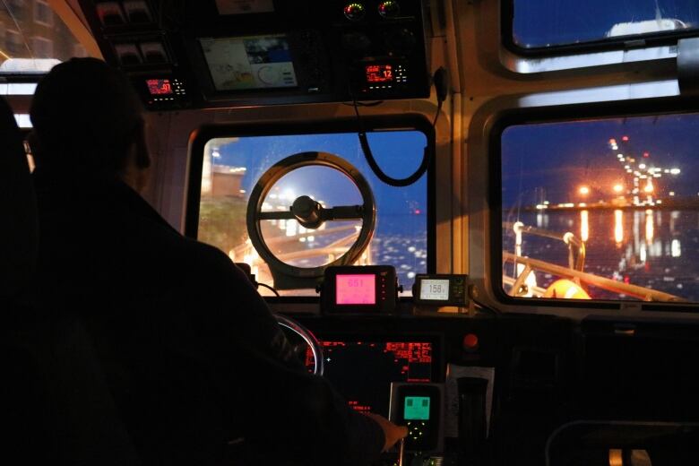 A darkened cockpit of a boat with city lights visible on either side of the Saint john harbour. 