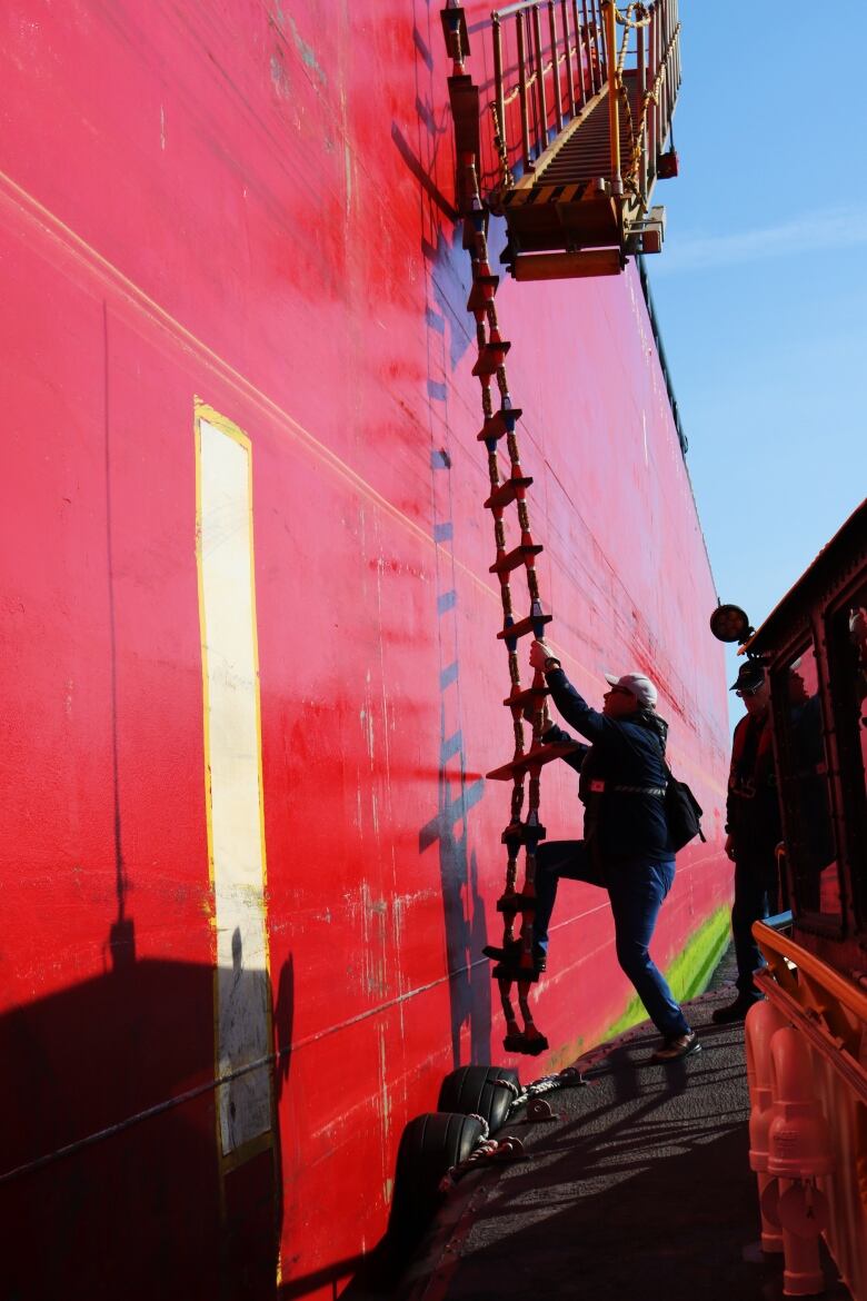 A harbour pilot starts his climb up a rope ladder down the side of a bright red Irving tanker. 