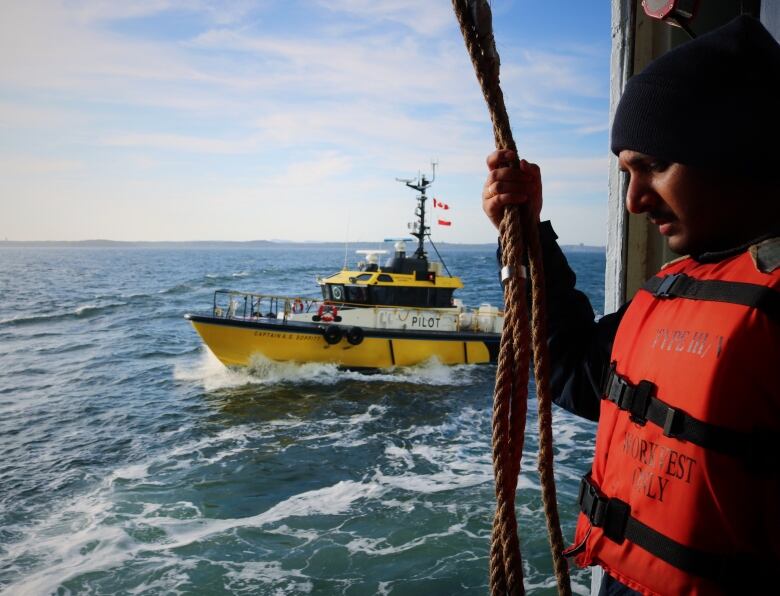 A pilot boat on choppy seas is visible through a hatch in the side of a cruise ship, which is held open by a young man wearing a black beanie. 