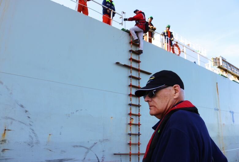 An older man watches a younger man climb a rope ladder down the side of a ship.