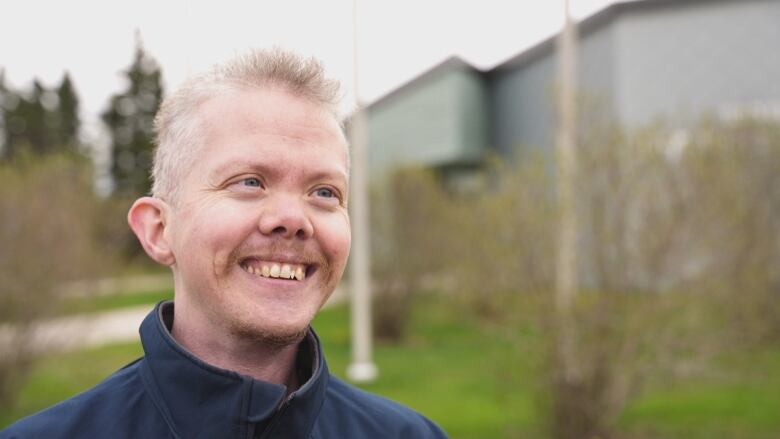 A man with short blond hair smiles in front of two flag poles.