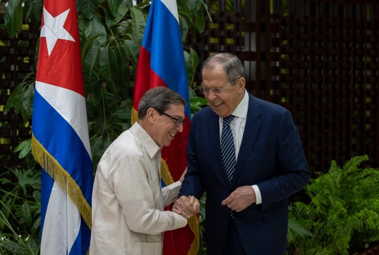 Two men shake hands and laugh in front of Russian and Cuban flags.