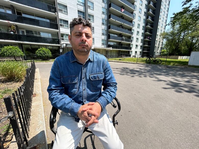 A man sits in a mobility aid in front of an apartment building. He is wearing a blue shirt and his hands are folded in front of him.