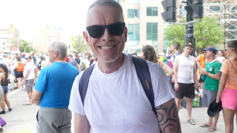 A man wearing sunglasses, a white T-shirt and a backpack is smiling at the camera.