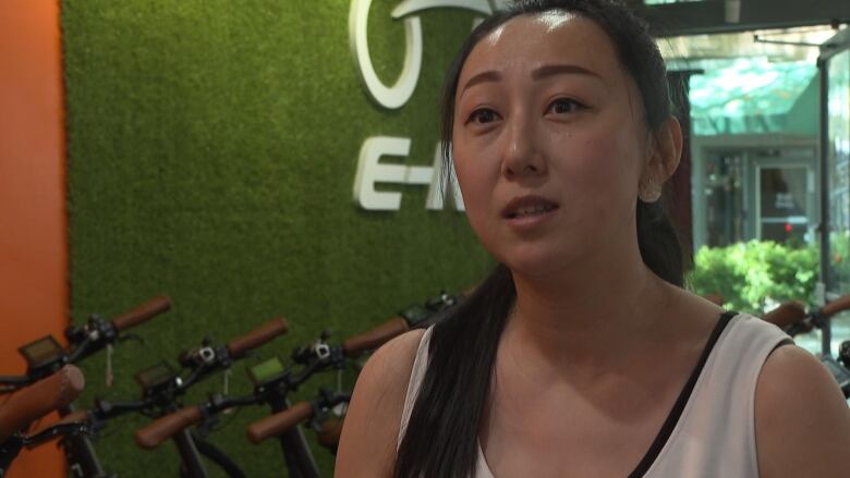 A woman speaks to the camera with a green wall and row of e-bikes behind her.