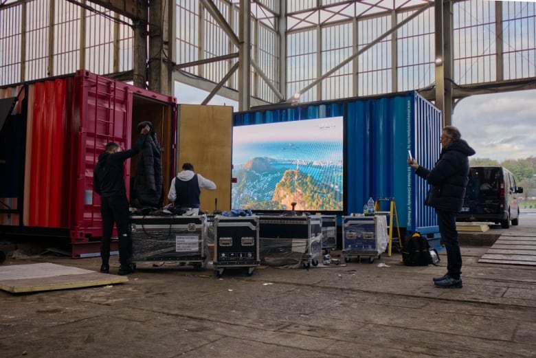 Two large shipping containers, one red and one blue, are set up in a warehouse space, with a screen displaying a mountain range out front. Three people unload gear out of one of the containers.