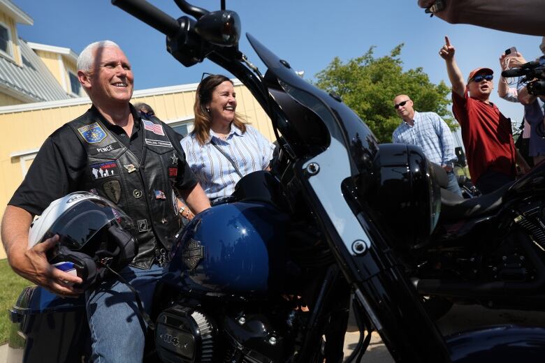 A white-haired man smiles while holding a helmet and sitting on a motorcycle.