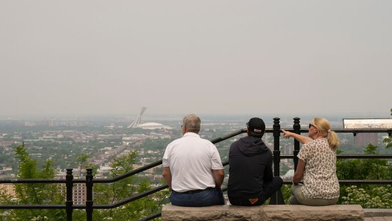 Three people seen from behind look at the skyline of Montreal, covered with a yellowish haze.