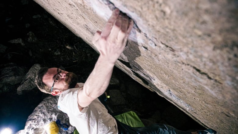 A shot from above of a bearded and bespectacled man in jeans and a T-shirt free-climbing a rock face, green padding on the ground beneath him.