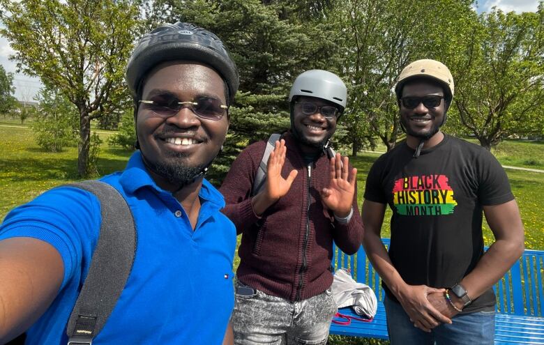 Three men take a selfie together in a park wearing bike helmets.