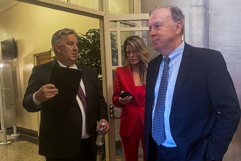 Two men in dark suits and a woman in a red suit stand in a hallway.