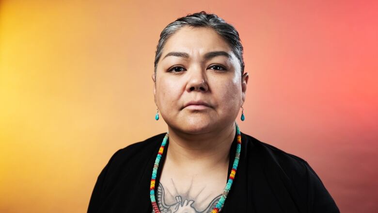 A woman wearing a beaded necklace looks serious in front of an orange background.