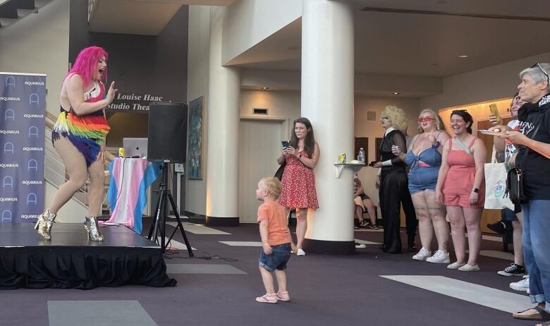 A drag performer waves to a small child, who is dancing along with her. 