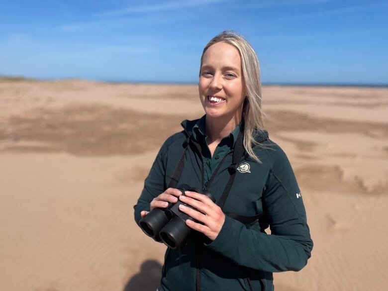 A woman in a Parks Canada uniform holding binoculars on a beach 