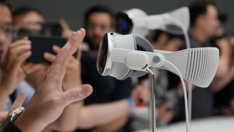 Two virtual reality headsets are displayed at a launch, as people take photos. In the foreground, a hand reaches toward a headset.