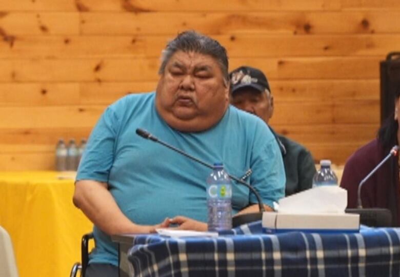 A man in a blue shirt sits in a wheelchair in front of a table with a microphone. A woman sits beside him. 