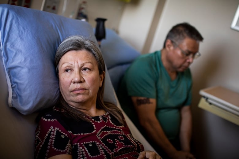 A woman lies in a hospital bed while a man sits next to her, head bowed.