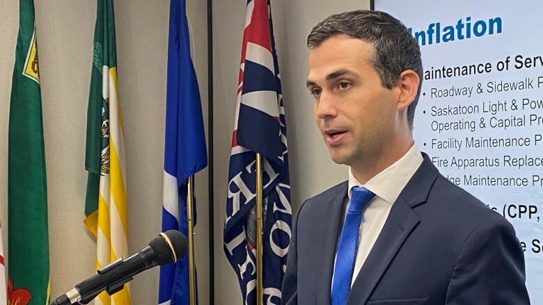 A man in a suit with a blue tie stands at a podium speaking into a microphone.